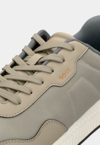 Gros plan sur une sneaker beige et grise avec un tissu texturé, un design à lacets et le logo doré "BOSS" sur le côté au-dessus d'une semelle blanche.