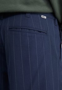 Pantalone s mornarskim prugama glatke teksture, s jednim stražnjim džepom i malom oznakom "OBEY" na pojasu. Jedva vidljive bijele pruge.