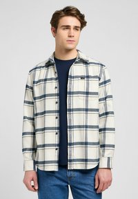 Camisa a cuadros de tonos claros con rayas azules y marinas, con botones en la parte delantera, un bolsillo en el pecho y un cuello plano, llevada sobre una camiseta oscura.