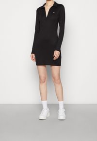 Black long-sleeve polo dress with a deep V-neck, slim fit, and subtle logo on the chest. Paired with white sneakers and socks.