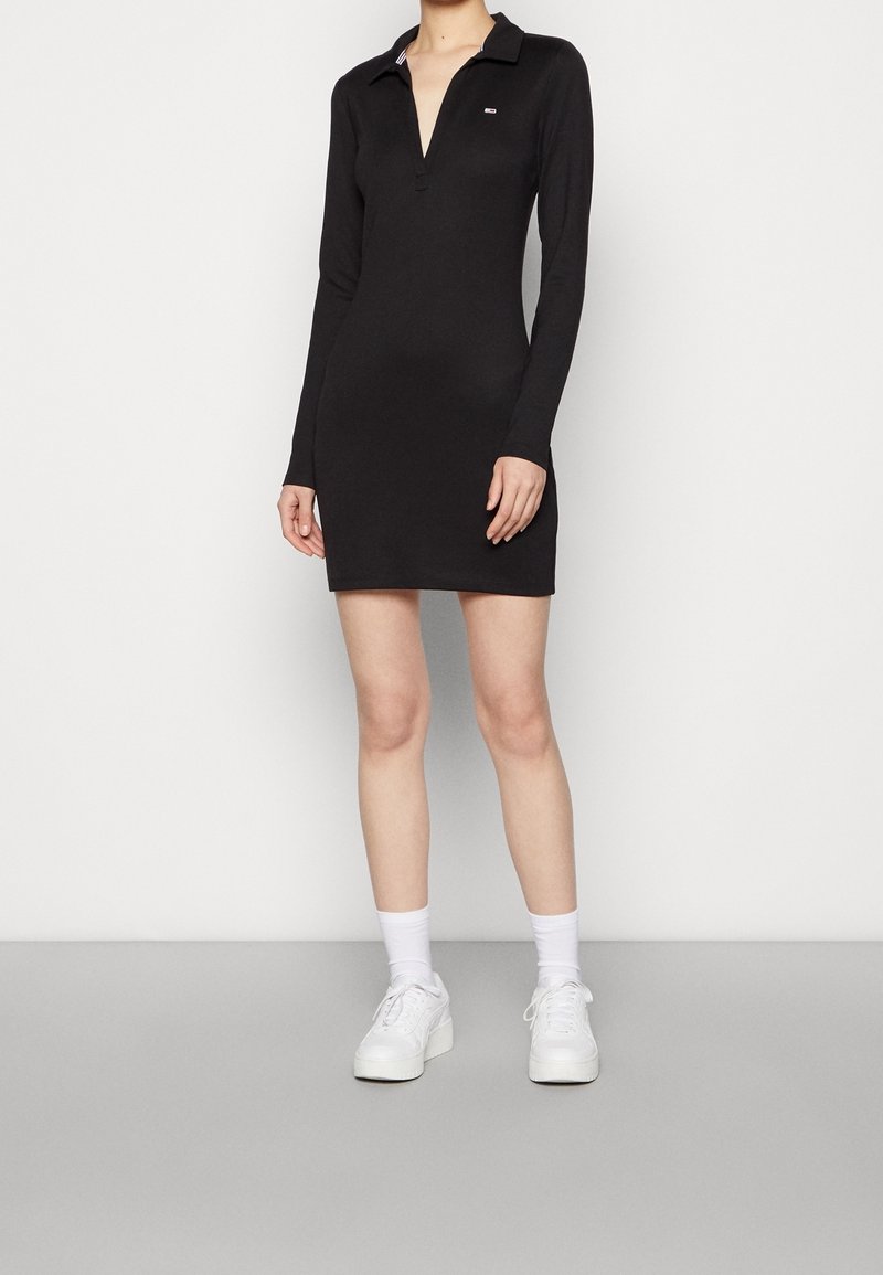 Black long-sleeve polo dress with a deep V-neck, slim fit, and subtle logo on the chest. Paired with white sneakers and socks.