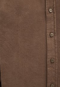 Braunes Cordhemd mit vertikaler Rippstruktur, ausgestattet mit einer Knopfleiste und runden Knöpfen mit eingravierter Markenbezeichnung.