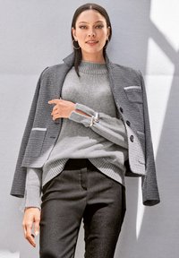 Schwarzer und weißer Hahnentrittmuster-Blazer mit zwei Knöpfen und Taschen, kombiniert mit einem hellgrauen gerippten Pullover und schwarzen maßgeschneiderten Hosen.