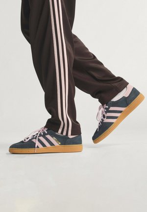 Zapatillas de ante navy con acentos rosas, que presentan tres rayas y cordones rosas, combinadas con pantalones deportivos marrones con rayas rosas.