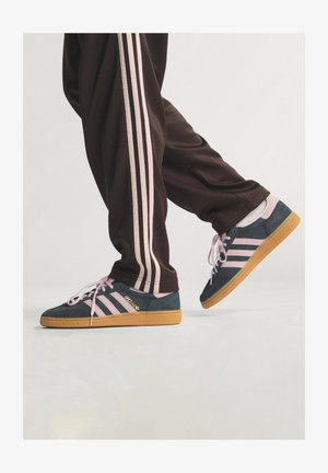 Granatowe zamszowe sneakersy z różowymi akcentami, z trzema paskami i różowymi sznurowadłami, w połączeniu z brązowymi spodniami sportowymi z różowymi paskami.