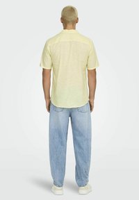 Lichtgeel kortemouwenshirt met een ronde zoom, gecombineerd met lichtblauwe, losse spijkerbroek en witte sneakers. Model bekeken vanaf de achterkant.