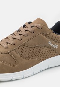 Light brown suede sneaker with perforated details, flat laces, and a white rubber sole. Features a textured surface and stitched accents.