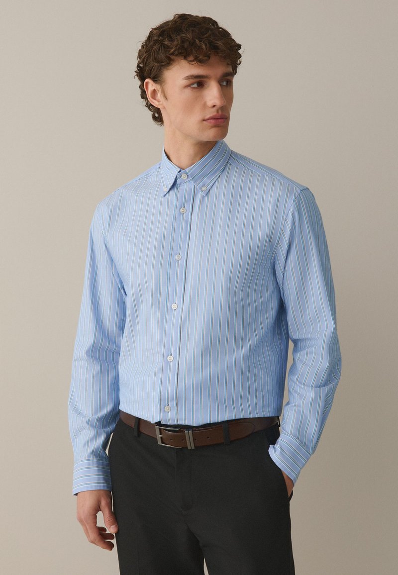 Joven con una camisa de botones de rayas azul claro metida en pantalones negros con un cinturón marrón, mirando hacia un lado.