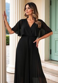 Czarna plisowana sukienka z przezroczystym, zwiewnym narzutem i dekoltem w kształcie litery V, charakteryzująca się miękkim materiałem i dopasowaną talią, na jasnym tle.