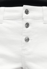 Άσπρο τζιν παντελόνι με τρία ορατά ασημένια μεταλλικά κουμπιά και θηλιές για ζώνη, φορεμένο με μαύρο τοπ.