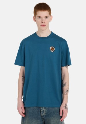 Giovane uomo con capelli rossi corti indossa una maglietta turchese con un piccolo logo circolare arancione e nero sul petto sinistro e jeans blu sbiaditi.