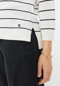 Pull blanc à rayures noires horizontales, présentant une fente sur le côté et un petit logo en métal. Associé à un pantalon noir et un bracelet en argent.