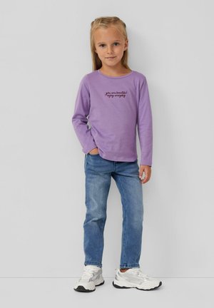 Paars t-shirt met lange mouwen met zwarte tekst "je bent mooi, geniet elke dag", gecombineerd met blauwe jeans en witte sneakers.
