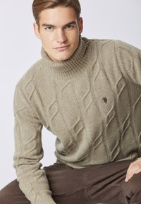 Maglione a collo alto beige lavorato a maglia con collo a coste, caratterizzato da un logo ricamato in modo discreto sul petto. Texture morbida, vestibilità rilassata.