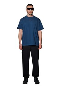 Marineblauwe katoenen t-shirt met "HALO" logo, gecombineerd met zwarte joggerbroeken met zijzakken en een trekkoord. Draagt een paar zwarte zonnenbril.