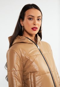 Hættepufferjakke i et blankt beige materiale. Har en fuld lynlås, sidelommer og et quiltet mønster.