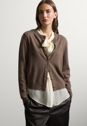 Mujer con un cárdigan marrón abotonado sobre una blusa blanca con un lazo, combinada con pantalones de cuero negros, de pie con las manos en los bolsillos.