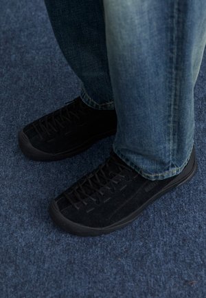 Black suede sneakers with textured surface, subtle lacing, and durable rubber soles, paired with blue denim jeans.