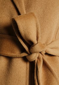 Gros plan sur un manteau en laine de couleur camel doux, noué en un large nœud lâche à la taille.