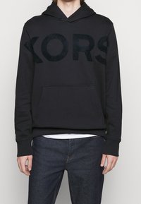 Sudadera con capucha negra que presenta un gran logo texturizado y oscuro "KORS" en la parte delantera, con un bolsillo tipo canguro y puños acanalados.