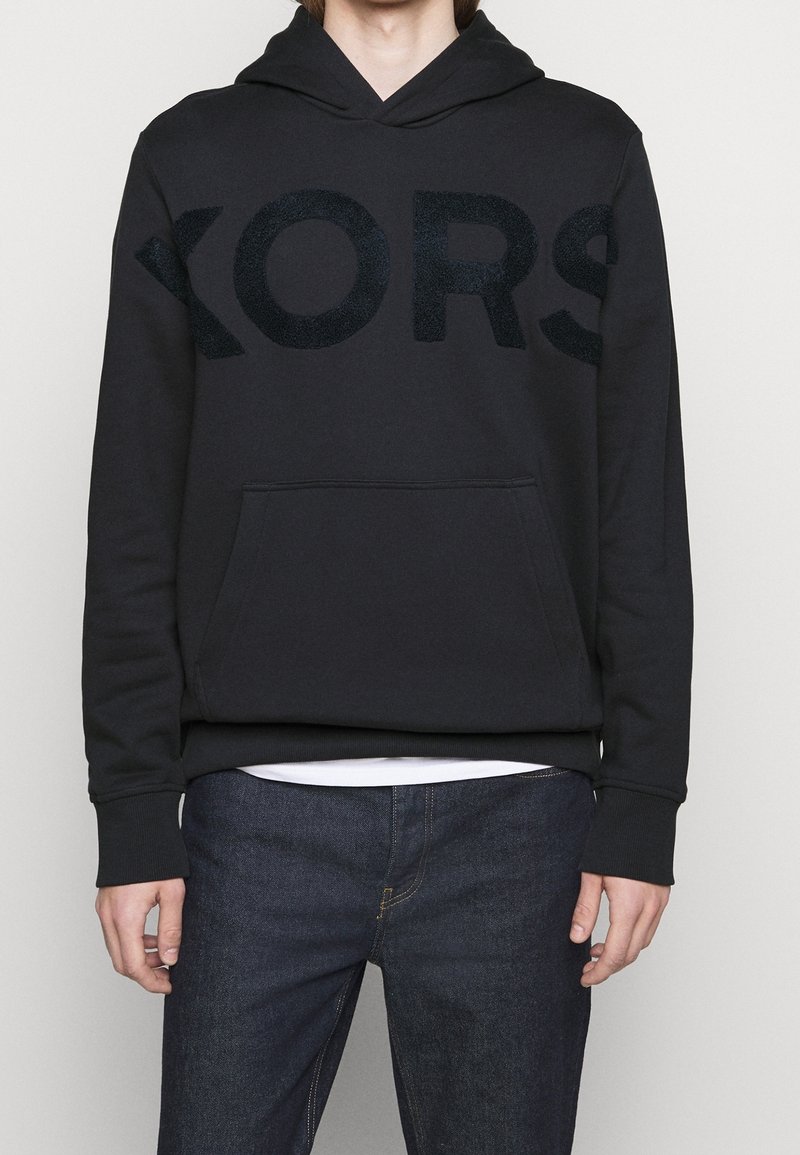 Sudadera con capucha negra que presenta un gran logo texturizado y oscuro "KORS" en la parte delantera, con un bolsillo tipo canguro y puños acanalados.