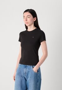 T-shirt nero aderente con maniche corte, scollo rotondo e piccolo logo ricamato in rosso, bianco e blu, abbinato a jeans in denim azzurro chiaro.