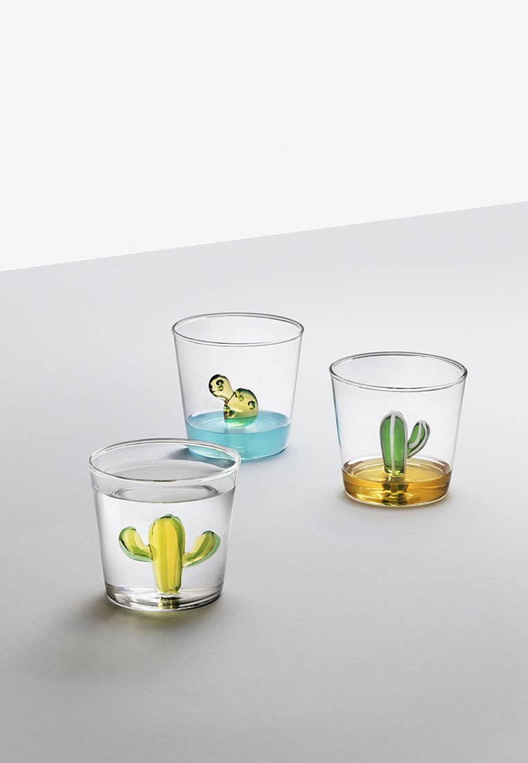 Tre bicchieri trasparenti con design di cactus colorati immersi in acqua colorata: blu, giallo e trasparente. Ogni bicchiere presenta dettagli di cactus verdi distintivi.