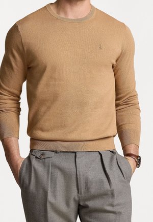 Mann trägt einen camel-farbenen Rundhalsausschnitt-Pullover mit einem kleinen eingestickten Logo, in graue, plissierte Hose gesteckt, die Hände in den Taschen.