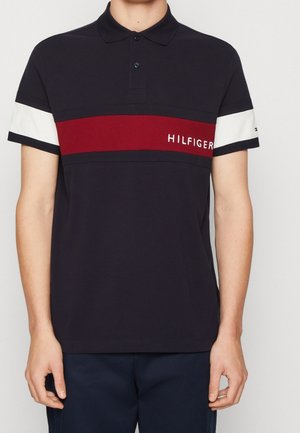 Navyblaues Poloshirt mit rotem Streifen und weißen Akzenten. Mit gesticktem "HILFIGER"-Logo. Kurze Ärmel, klassischer Kragen und normale Passform.
