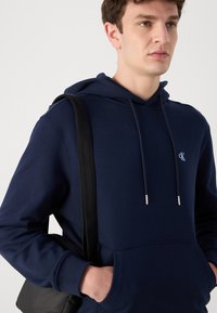Felpa in cotone blu navy con tasca frontale, cappuccio regolabile con cordino e piccoli occhielli metallici. Logo piccolo sul petto, indossata con una borsa nera.