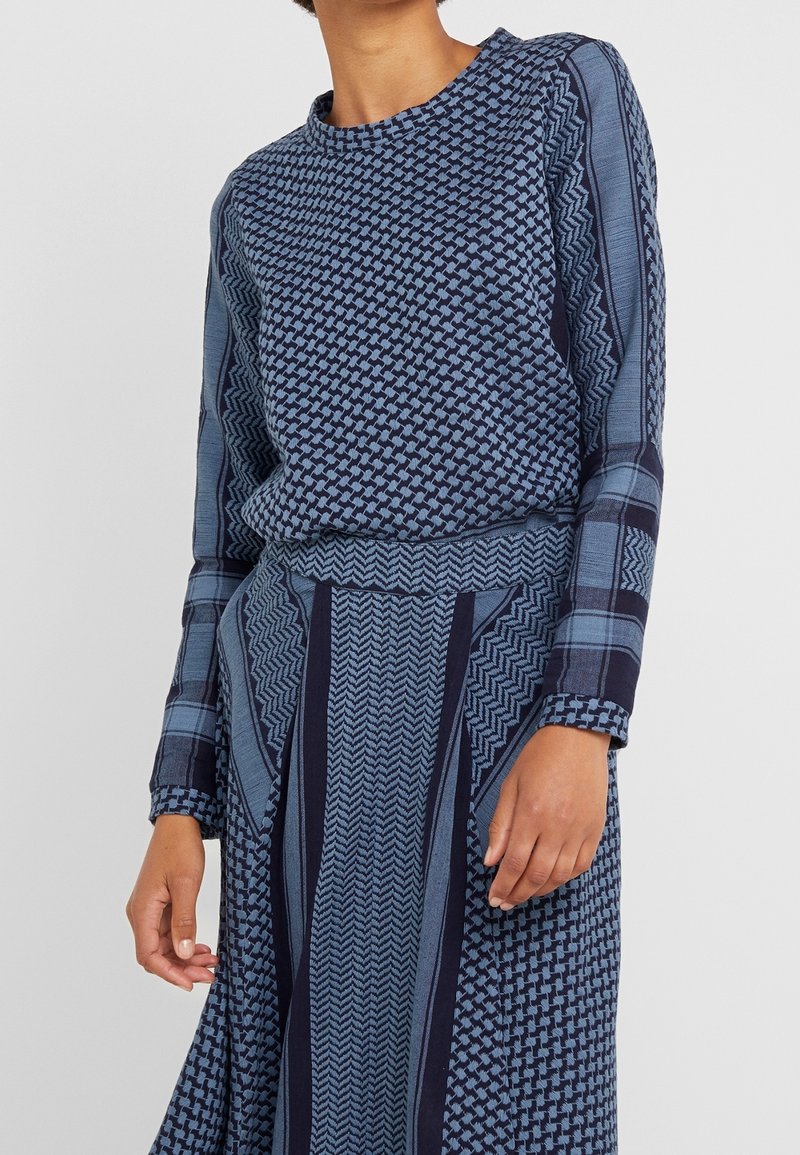 Femme portant un haut bleu à manches longues à motifs et une jupe aux dessins géométriques et à carreaux, debout devant un fond clair uni.