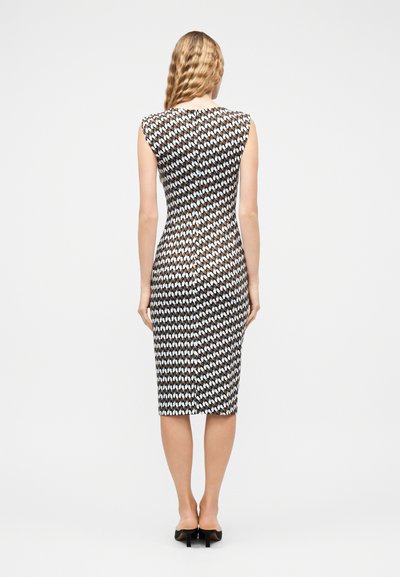 Γυναίκα με μακριά κυματιστά ξανθά μαλλιά, φορώντας αμάνικο, μίντι φόρεμα με σχέδια και μαύρα παπούτσια με τακούνι, όρθια με την πλάτη προς τα εμπρός.