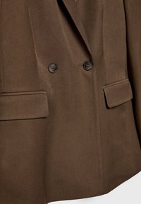 Blazer marron croisé avec deux boutons noirs, deux poches avant et une texture de tissu lisse. Coupe ajustée et design minimaliste.