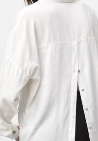 Femme portant une chemise blanche à manches longues avec une ouverture dans le dos fermée par de petits boutons, révélant des vêtements noirs en dessous.