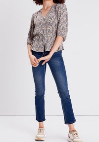 Blusa floreale con scollatura a V, maniche a tre quarti e vita a peplo, abbinata a jeans in denim scuro e sneakers beige.