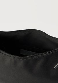 Mustast kangast kotike, millel on osaliselt avatav lukk, paljastades tumeda sisu ja sees Lacoste'i silt.