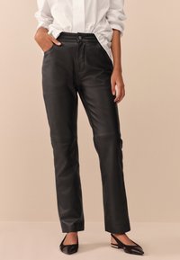Pantalon en cuir noir à la texture lisse, doté de poches avant, d'une fermeture par bouton et d'une silhouette droite. Des chaussures plates noires complètent le look.