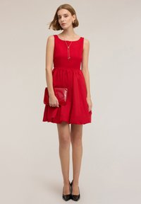 Punainen hihaton mekko, jossa on vartalonmyötäinen yläosa ja levenevä helma. Malli pitää käsissään kiiltävää punaista clutch-laukkua, joka on yhdistetty mustiin teräväkärkisiin korkokenkiin.