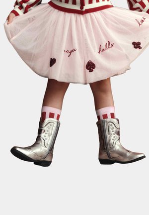 Bambina che indossa una gonna di tulle rosa con parole e simboli ricamati, calzini a righe e stivali metallici argento, in piedi davanti a uno sfondo semplice.
