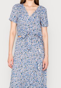 Haut court à fleurs bleu avec manches courtes et détail noué à l'avant, associé à une jupe assortie présentant un motif floral similaire et des volants.