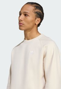 Beige sweatshirt med rund halsringning, raglanärmar och en liten vit Adidas-logotyp på bröstet. Slät textur, minimalistisk design.