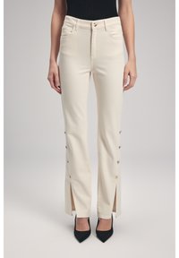 Pantaloni beige chiaro a vita alta con aperture laterali fissate da bottoni dorati, dotati di tasche frontali e design aderente.