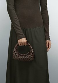 Brun vävd läderhandväska med ett runt handtag, texturerad yta och kompakt form. Modellen bär en ribbad brun topp och en mörkgrön kjol.