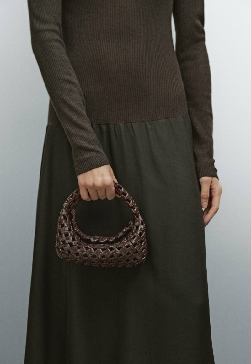 Brun vävd läderhandväska med ett runt handtag, texturerad yta och kompakt form. Modellen bär en ribbad brun topp och en mörkgrön kjol.