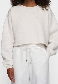 Donna che indossa un crop sweatshirt color crema con il sottile testo "10 DAYS" e pantaloni della tuta chiari con cordoncino abbinato, mano in tasca.