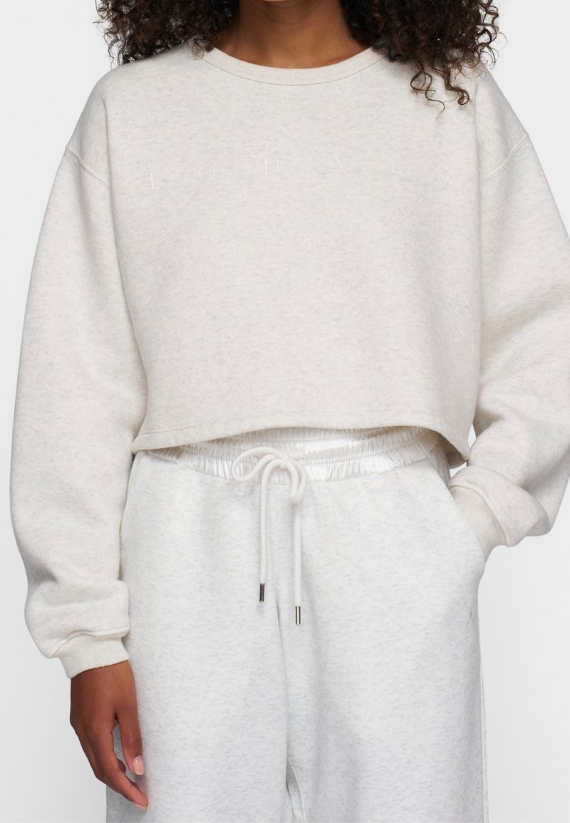 Donna che indossa un crop sweatshirt color crema con il sottile testo "10 DAYS" e pantaloni della tuta chiari con cordoncino abbinato, mano in tasca.