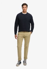 Marineblauwe gebreide trui met textuur, beige broek en blauwe sneakers. De trui heeft een ronde hals; de broek is met opgerolde zoom. Casual outfit.