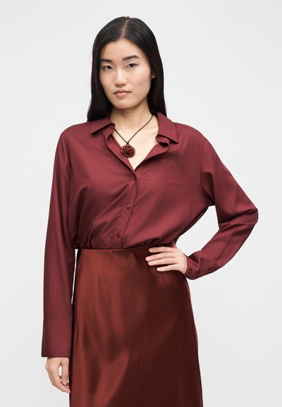 Bordowa bluzka z kołnierzykiem, długimi rękawami i zapięciem na guziki. Połączona z dopasowaną satynową spódnicą. Prosty naszyjnik z kwiatowym motywem.