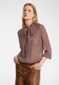 Blusa trasparente marrone e crema con colletto a fiocco, caratterizzata da un motivo geometrico di punti, vestibilità ampia e maniche a tre quarti.