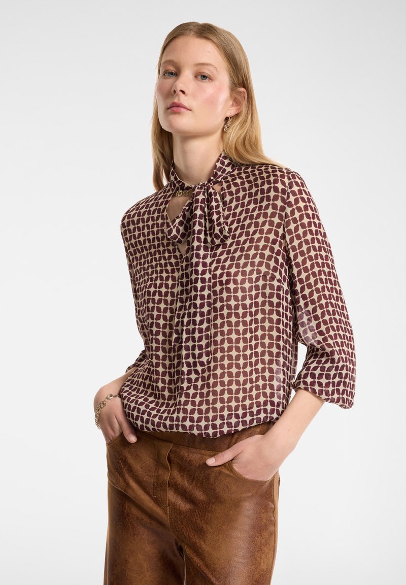 Blusa trasparente marrone e crema con colletto a fiocco, caratterizzata da un motivo geometrico di punti, vestibilità ampia e maniche a tre quarti.
